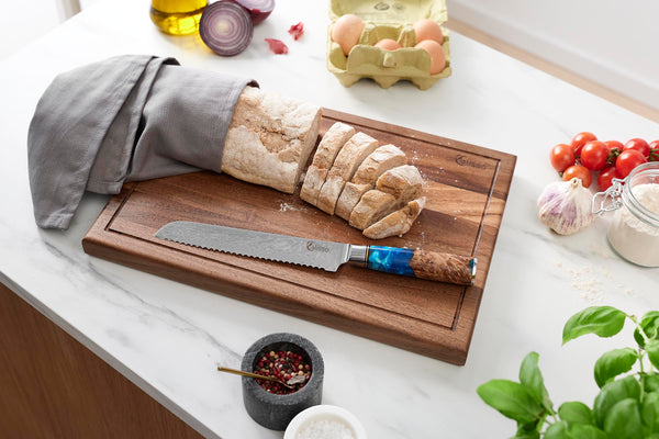 Calisso Brotmesser neben frisch gebackenem Baguette, eingewickelt in ein graues Tuch, mit Öl, Eiern, Kräutern und Gewürzen im Hintergrund.